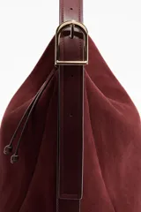 Cartera de hombro de gamuza color bordó, con cierre superior y tirador con detalles de nudos. Cuenta con correa ajustable mediante hebilla metálica en los laterales y diseño estructurado pero flexible.