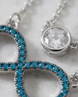 Collar doble de plata 925, compuesto por una cadena superior con dije de punto de luz y una cadena inferior con dije en forma de símbolo de infinito decorado con circonias turquesas.