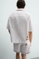 Camisa de corte relajado confeccionada en algodón, con cuello bowling y manga corta. Presenta un bolsillo en el pecho con vivo a contraste y bordados decorativos en el frente. Cierre frontal mediante botones.