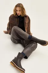 Bota de caña media confeccionada en cuero vacuno texturizado color marrón oscuro. Presenta un diseño minimalista con costuras reforzadas, tirador trasero y una suela de plataforma plana con capas en tonos beige, negro y suela.