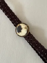 Cinto trenzado de cuero color marrón oscuro con hebilla circular decorativa que presenta un inserto de piedra natural con vetas en tonos crema, marrón y negro, enmarcada en metal dorado.