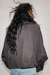 Chaqueta estilo bomber de color gris oscuro, con cuello subido, manga larga y corte holgado. Presenta bolsillos delanteros con solapa, bajo con pliegues y cierre frontal con cremallera oculta y botones a presión.