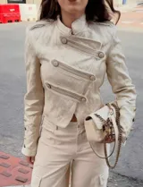 Chaqueta de estilo militar confeccionada en tejido jacquard color beige con sutil brillo. Presenta cuello mao, cierre frontal cruzado con botones decorativos, hombreras con detalles de botones y puños con botones. Posee un calce entallado y largo a la cintura.