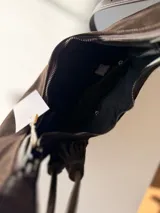 Cartera tipo hobo de textura gamuzada en color marrón oscuro, con diseño flexible y asa larga regulable. Presenta un detalle de flecos largos desmontables en uno de los laterales y cierre superior.