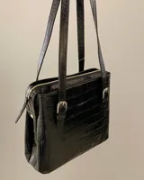Cartera de hombro estructurada color negro, confeccionada en 100% cuero vacuno con textura en relieve tipo reptil. Cuenta con asas largas ajustables mediante hebillas metálicas plateadas, cierre superior y el logo de la marca grabado en la parte frontal.