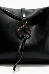 Cartera bandolera de hombro en color negro, confeccionada en material sintético con acabado liso. Presenta una solapa frontal con un detalle metálico circular decorativo y cierre magnético oculto. Incluye una correa ajustable y un compartimento interior con cierre.