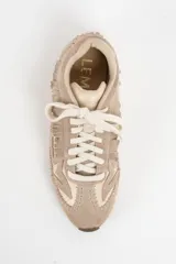 Zapatilla de cuero legítimo en tonos beige y crema, con diseño de flecos en el lateral y costuras decorativas visibles en el contorno. Presenta cordones al tono y suela plana.