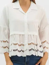 Camisa blanca de corte recto con cuello camisero y cierre frontal de botones. Presenta mangas tres cuartos acampanadas con detalles de bordado suizo calado en los puños y en el ruedo inferior.