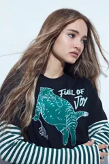 Remera negra de manga corta con estampa gráfica de un gato verde y la frase 'Full of Joy'. Presenta un diseño de doble manga con una capa inferior de rayas horizontales blancas y verdes.
