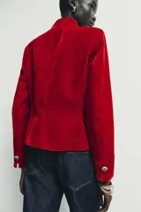 Blazer de terciopelo color rojo, con cuello subido, manga larga y cierre frontal cruzado con botones dorados. Presenta bolsillos delanteros con solapa.
