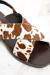 Sandalias bajas con tiras cruzadas de cuero con estampado de vaca en tonos marrón y blanco. Tira trasera con hebilla dorada y suela plana color marrón.