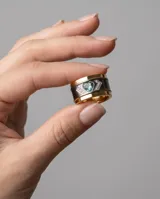 Anillo ancho de acero con banda central oscura, bordes con baño de oro, engastado con una circonia en forma de corazón de color celeste y detalles de pequeñas piedras brillantes.