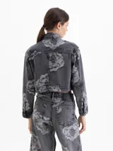 Chaqueta cropped de denim con lavado gris y estampado floral en contraste. Presenta cuello clásico, mangas largas, bolsillos frontales con tapa y cierre mediante broches metálicos.