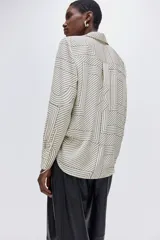 Camisa de manga larga con cuello camisero y cierre frontal de botones. Presenta un estampado geométrico en blanco roto y negro, compuesto por líneas rectas y curvas que forman patrones abstractos.