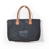 Cartera tipo tote de color negro, combinada con asas y detalles en símil cuero color tostado. Cuenta con una pequeña placa metálica con el logo de la marca en el frente.