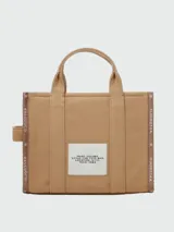 Bolso tipo tote bag de Marc Jacobs en color beige, confeccionado en lona resistente. Presenta el texto "THE TOTE BAG" y "MARC JACOBS" impreso en la parte frontal en color negro. Cuenta con asas superiores y una correa de hombro ajustable y extraíble, además de ribetes laterales negros con inscripciones.