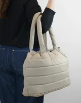 Bolso tipo tote color beige con diseño acolchado y asas tubulares. Presenta el logo de la marca en el centro.