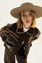 Campera de lana merino hervida en color marrón, con diseño de corte recto, cuello camisero y cierre frontal con botones. Presenta detalles de bordados artesanales en contraste con motivos geométricos y líneas onduladas en los bolsillos frontales y solapas.