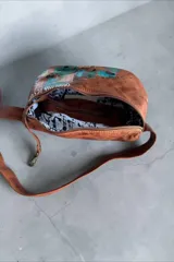 Riñonera de cuero terracota con diseño pintado a mano en tonos turquesa y dorado, con cierre superior y correa ajustable con herrajes metálicos.