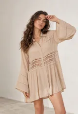 Vestido corto beige estilo camisa con mangas largas amplias, cuello camisero, cierre frontal con botones y detalles de encaje calado en el pecho y las mangas.