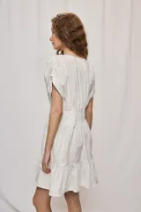 Vestido corto de algodón color blanco, con escote en V y botonadura frontal. Presenta mangas cortas holgadas, cintura ajustada con frunces y falda acampanada con terminación de volado en el ruedo.
