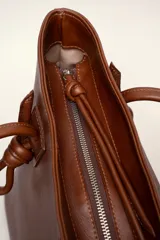 Cartera tote de cuero suave color marrón, con diseño amplio y líneas simples, con tiras de hombro decoradas con nudos artesanales y pequeño logo grabado en el centro.