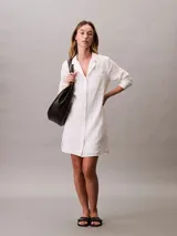 Vestido camisero corto de color blanco, confeccionado en tejido de gasa de algodón. Presenta cuello clásico, cierre frontal con botones, mangas largas y un bolsillo aplicado en el pecho.