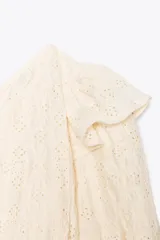 Top corto asimétrico color blanco roto, con un solo hombro descubierto y manga abullonada corta en el otro lado. Presenta bordados perforados (ojales) y un volante en el hombro descubierto. El bajo y el puño de la manga terminan en elástico.