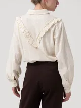 Blusa de algodón orgánico color crema con cuello camisero, cierre frontal de botones y mangas largas con puños. Presenta un diseño con volantes bordados que recorren el frente y detalles de bordado calado a lo largo de toda la prenda.