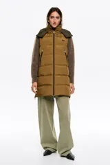 Chaleco acolchado de largo medio y corte en línea A, confeccionado en tejido técnico color tostado. Presenta cuello subido con capucha desmontable, cierre frontal de cremallera, bolsillos laterales con cremallera, logo bordado en el pecho y aberturas laterales con cremallera.