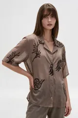 Camisa de algodón marrón con estampado abstracto oscuro, de corte holgado, mangas cortas y cuello solapa con cierre frontal de botones.