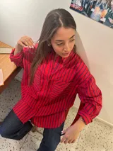 Camisa de corte oversize con diseño de rayas verticales en color rojo y azul. Presenta cuello clásico, cierre frontal con botones, mangas largas con puños abotonados y dos bolsillos frontales con solapa. El diseño tiene un largo asimétrico, siendo más corto en la parte delantera y más largo en la parte trasera.