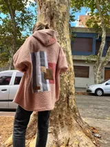Campera de corderito sintético color rosa, con capucha y diseño de collage en la espalda que combina franjas verticales en tonos grises, blanco y parches en texturas de marrón y terracota. Presenta un corte holgado y mangas anchas.