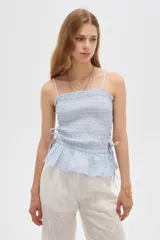 Top de algodón con diseño estampado de cuadros vichy en tonos celeste y blanco, corte al cuerpo con escote recto y tirantes finos. Presenta cuerpo fruncido tipo smock con cordones ajustables en los laterales que forman un pequeño volado en el ruedo.