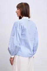 Blusa celeste con rayas verticales, cuello en V, mangas largas abullonadas con puños blancos y volantes verticales en el frente.