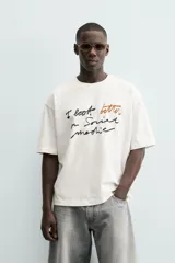 Camiseta blanca de algodón con corte boxy fit y cuello redondo. Presenta un estampado frontal con texto manuscrito en negro y marrón.