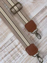 Correa de repuesto para cartera, de tela color beige con rayas blancas y negras. Incluye herrajes metálicos color bronce y apliques de cuero sintético color marrón.