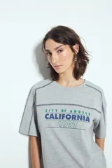 Remera gris de manga corta con estampado tipográfico frontal que dice "CITY OF ANGELS CALIFORNIA 1997" en colores verde y azul. Presenta costuras visibles en color azul oscuro que recorren los hombros y los laterales.