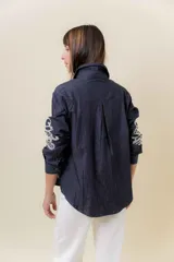 Camisa de denim liviano color azul oscuro con bordado artesanal en tono crudo en el frente y en las mangas. Presenta cuello clásico y cierre frontal con botones, con un calce relajado.