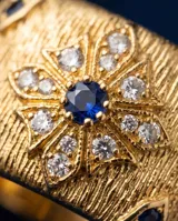 Anillo ancho de bronce con baño de oro, acabado texturizado y diseño con flor central incrustada de circonias blancas y una circonia azul en el centro.