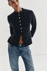 Chaqueta de punto color negro con cuello redondo, manga larga y cierre frontal con botones dorados. Presenta un corte entallado con bajo en evasé.