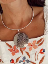Gargantilla rígida en tono plateado con un dije circular grande de superficie texturizada con diseño de puntos concéntricos.