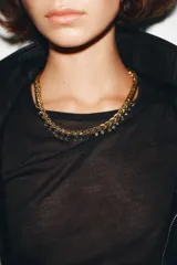 Collar tipo gargantilla de eslabones trenzados en tono dorado, con pequeños colgantes en forma de pirámide o cono en tono oscuro (negro/gris) colgando a lo largo de la parte inferior. Posee un cierre de caja cuadrado en la parte trasera.