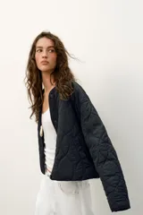 Cazadora corta acolchada de color negro, con cuello redondo, manga larga y cierre frontal con botones. Presenta un diseño de costuras geométricas y bolsillos laterales de vivo.