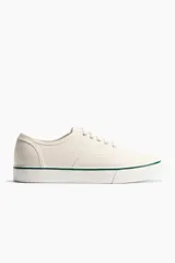 Calzado deportivo de lona de algodón color crudo, con cordones y suela de goma blanca con una línea verde.