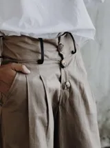 Pantalón ancho de tiro alto color beige, con pliegues frontales y botones visibles en la cintura. El ruedo del pantalón tiene un detalle de terminación en cuero color caramelo.