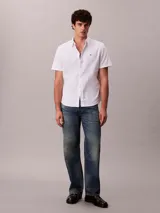 Camisa blanca de manga corta, confeccionada en tejido Oxford de algodón orgánico con elastano. Presenta cierre frontal con botones, cuello abotonado y logo de Calvin Klein Jeans bordado en el pecho.