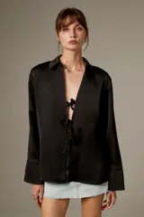 Blusa negra de poliéster, con cuello camisero, mangas largas y cierre frontal mediante tiras para anudar.