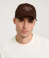 Gorra de sarga de algodón color marrón oscuro con visera curva y ajuste trasero. Presenta un bordado frontal con las letras 'NYC' en estilo universitario con detalles en rojo y blanco, flanqueadas por los números '19' y '86' y tres estrellas blancas debajo.