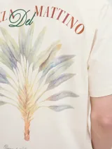 Remera color hueso de algodón con cuello redondo y manga corta. Estampado en la espalda con diseño de hojas de palmera y texto "Brezza Del Mattino Originals Summer Edition EST 1990".